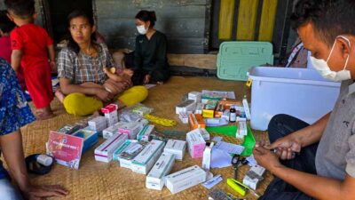 People sit on a woven mat sorting and distributing assorted medication boxes in a relief setting, with masks on some individuals.
