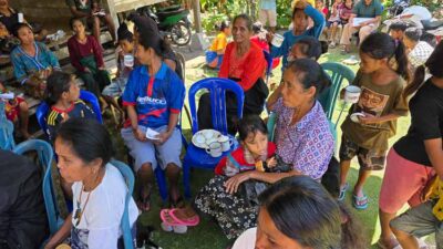 Group of women and children sitting on plastic chairs outdoors at a community gathering, with cups and plates nearby.