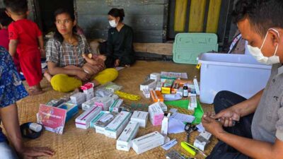 People sit on a woven mat sorting and organizing boxes of medicine and medical supplies, with masks on in a community setting.