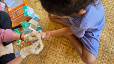 Healthcare worker in gloves applies a bandage to a child's finger on a woven mat, with medical supplies nearby.