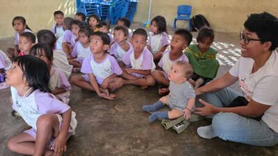 Group of young children in purple-and-white uniforms sit on the floor in a circle, listening to a man with glasses on the right in a classroom setting.