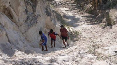 Three children climb up a rocky, dusty path carved into a white cliffside, using handholds and stepping carefully on uneven terrain.