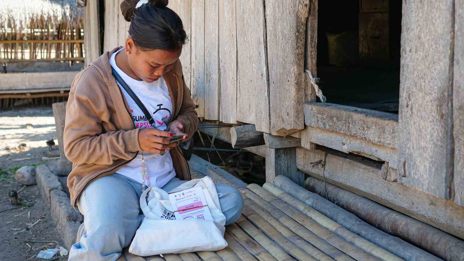 Rambu Fratika using a phone for Sumba Sepsis Study fieldwork beside a wooden house in East Sumba.