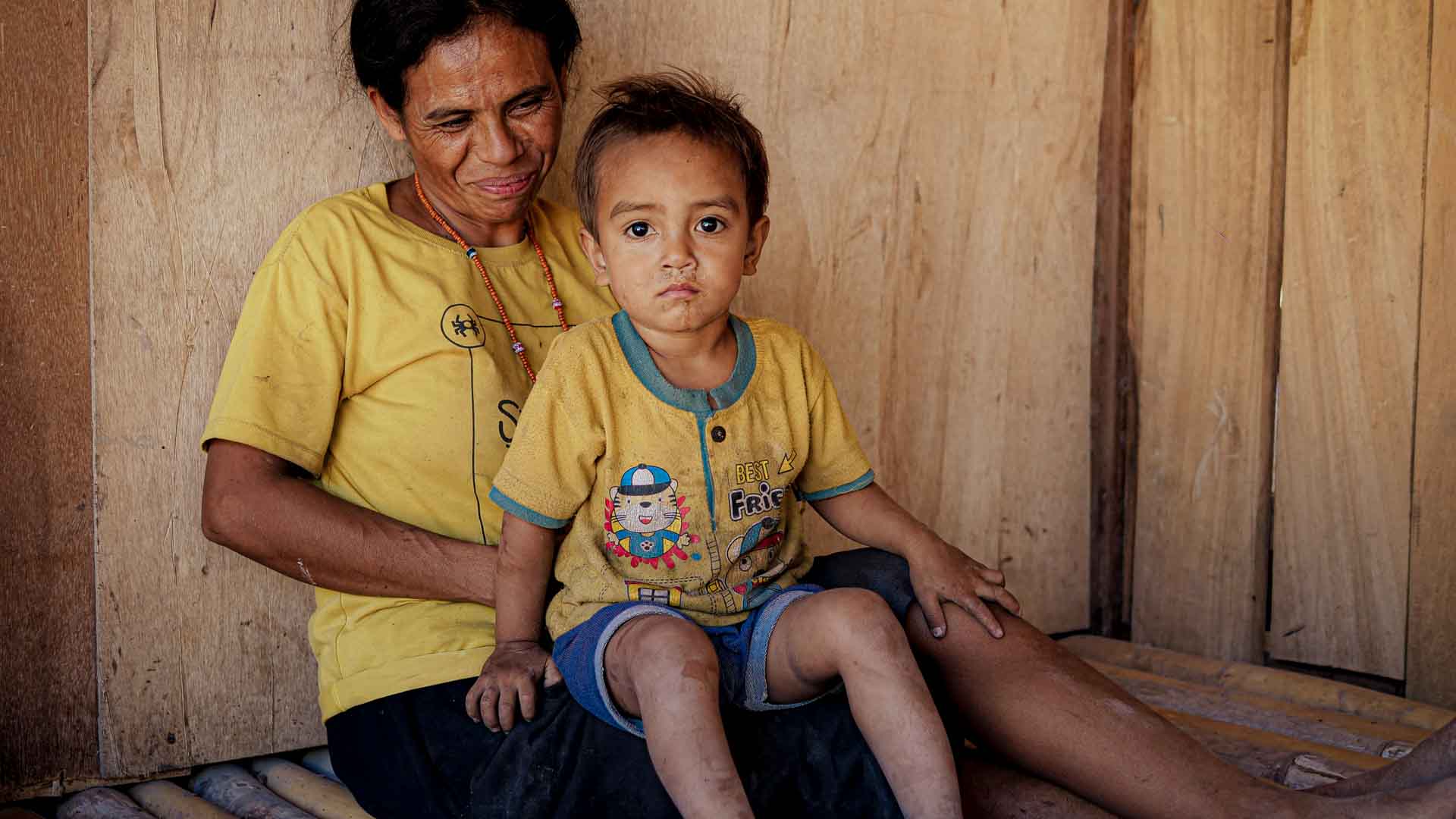 Rural Child Health in East Sumba Mother in a yellow shirt holding a young child on her lap beside a wooden wall in Napu, East Sumba.
