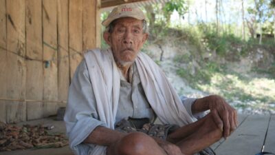 Un uomo più anziano con una casacca seduto fuori da una casa di legno a Mbajik, East Sumba, guardando verso la telecamera.