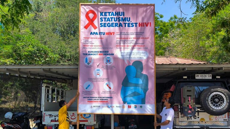 Two team members holding a large Indonesian HIV testing billboard outdoors beside field vehicles at Rumah Kambera.