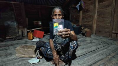 Older woman seated in a wooden home holding medicines received during a Kawan Sehat primary healthcare visit.