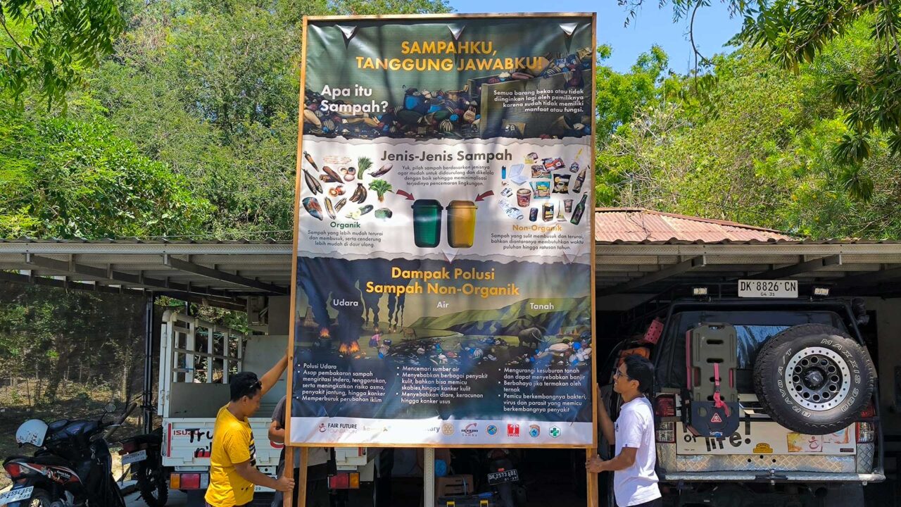 waste sorting billboard at rumah kambera before installation Two team members holding a tall Indonesian waste sorting billboard beside field vehicles at Rumah Kambera.