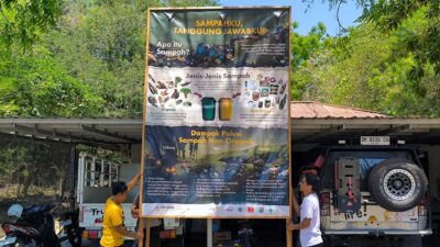 Two team members holding a tall Indonesian waste sorting billboard beside field vehicles at Rumah Kambera.