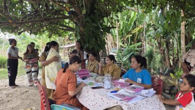 Mothers, children and health workers around records at an outdoor Posyandu table in Lambanapu, East Sumba.