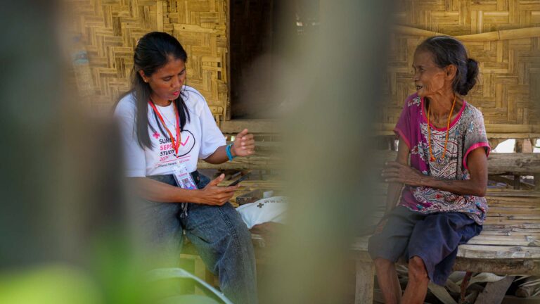 Un agent de Sumba Sepsis Study terrain parle avec une femme âgée devant une maison en bambou tissé.