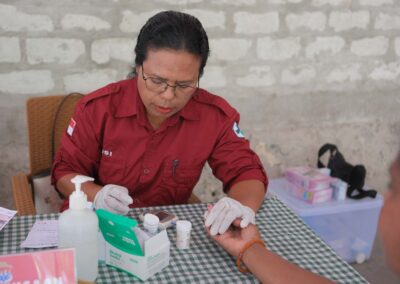 A gloved health worker holds a resident’s fingertip beside a portable meter and health cards.