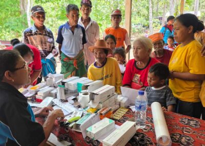 A health worker speaks with children and adults gathered around a table of medicines in Bidi Hunga.