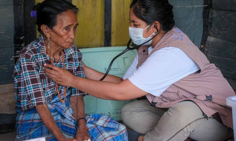 Un employé de santé masqué utilise un stéthoscope pour examiner une femme assise.