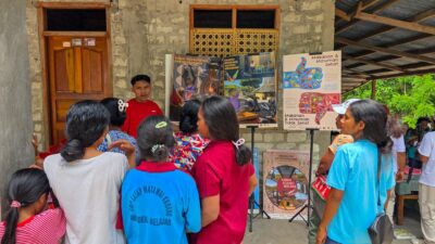 Erwin in red speaks to a group beside tobacco, alcohol and nutrition posters, with a malaria poster below.