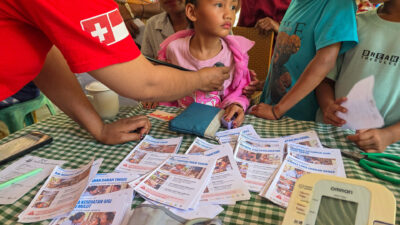 A doctor examines a child in pink with a stethoscope beside health cards; Swiss and Indonesian flags mark the sleeve.