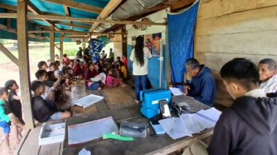 Desiana leads health education beside fabric posters as children and adults sit nearby in Lahiru.