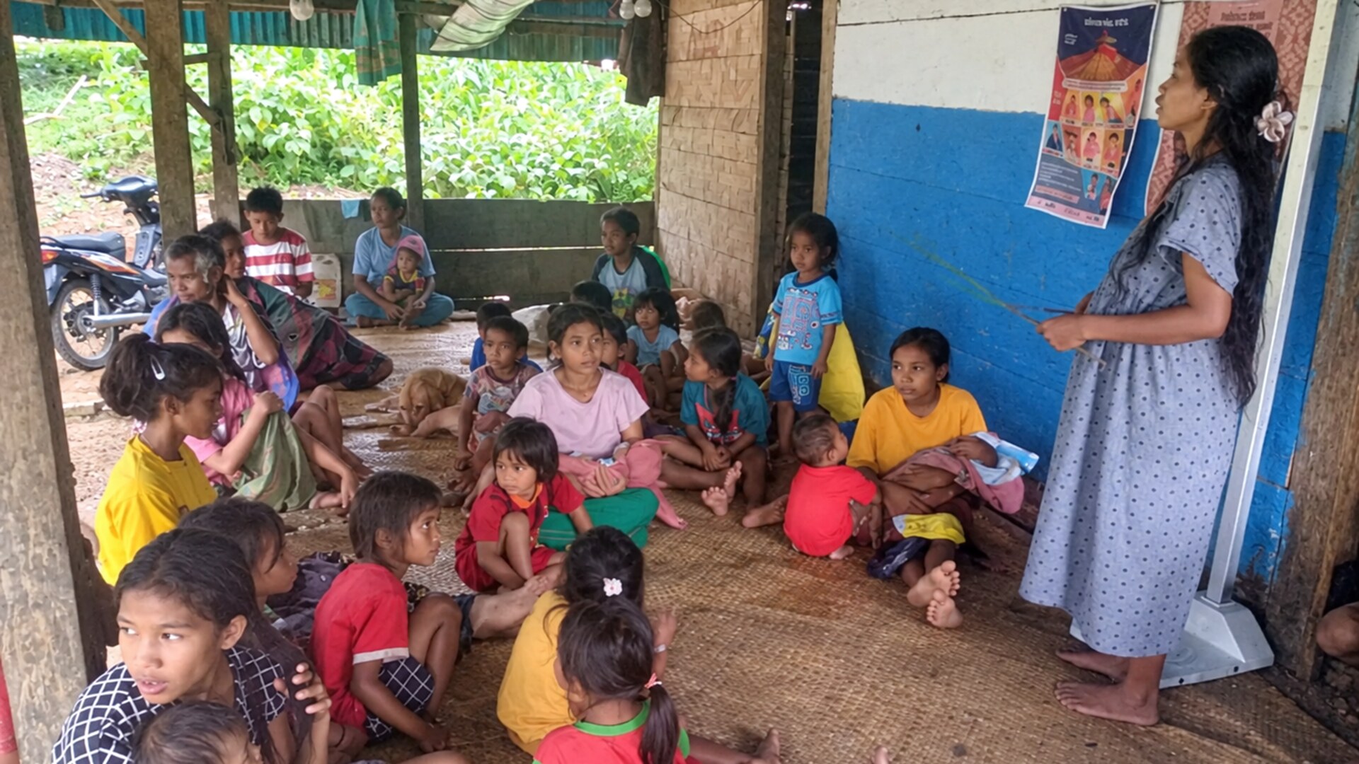 Children and adults on woven mats beside a woman using a pointer and health education posters in Laihiru.