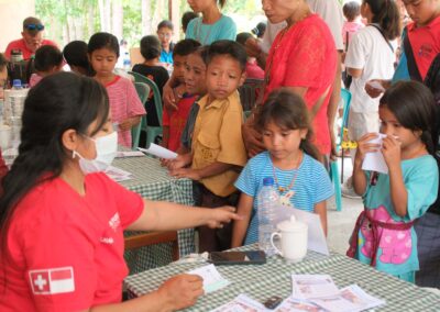 A masked health worker reaches towards a girl's arm, with children, adults and illustrated health cards around the consultation table.