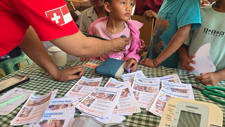 A healthcare worker examines a child with a stethoscope in Bidi Hunga, East Sumba, while illustrated health prescription cards are displayed on the table.