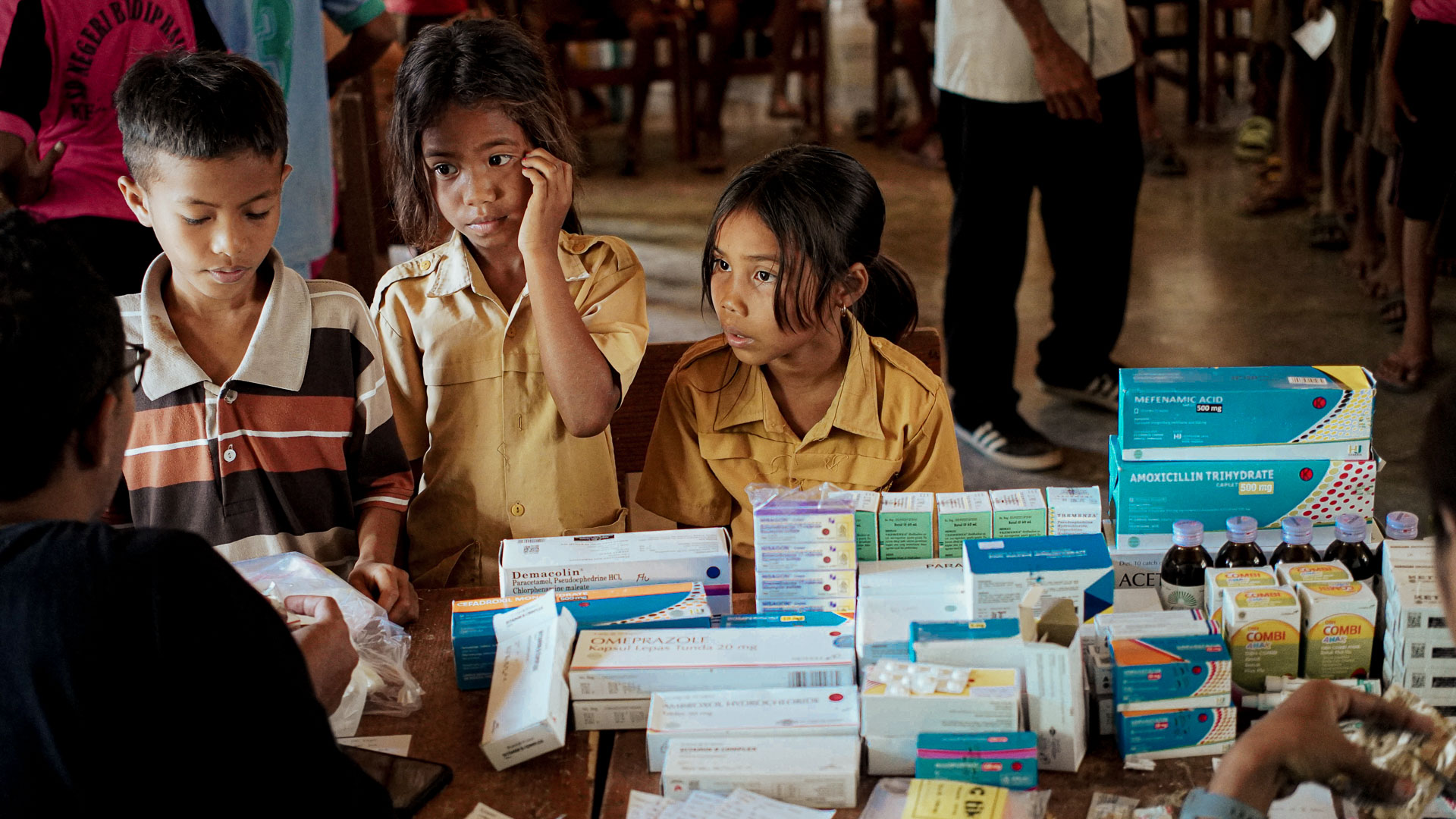 Medicines at a community care day Three children face an adult across a table of medicines, with other people and wooden seating behind them.