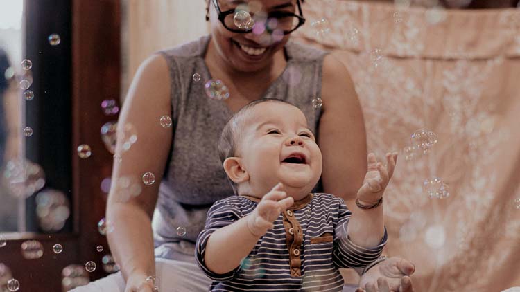 Baby Sorai laughs and reaches for floating soap bubbles while his mother Ayu smiles behind him.