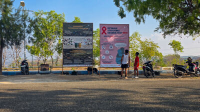 People stand beside HIV and waste billboards near parked scooters and trees in Waingapu.