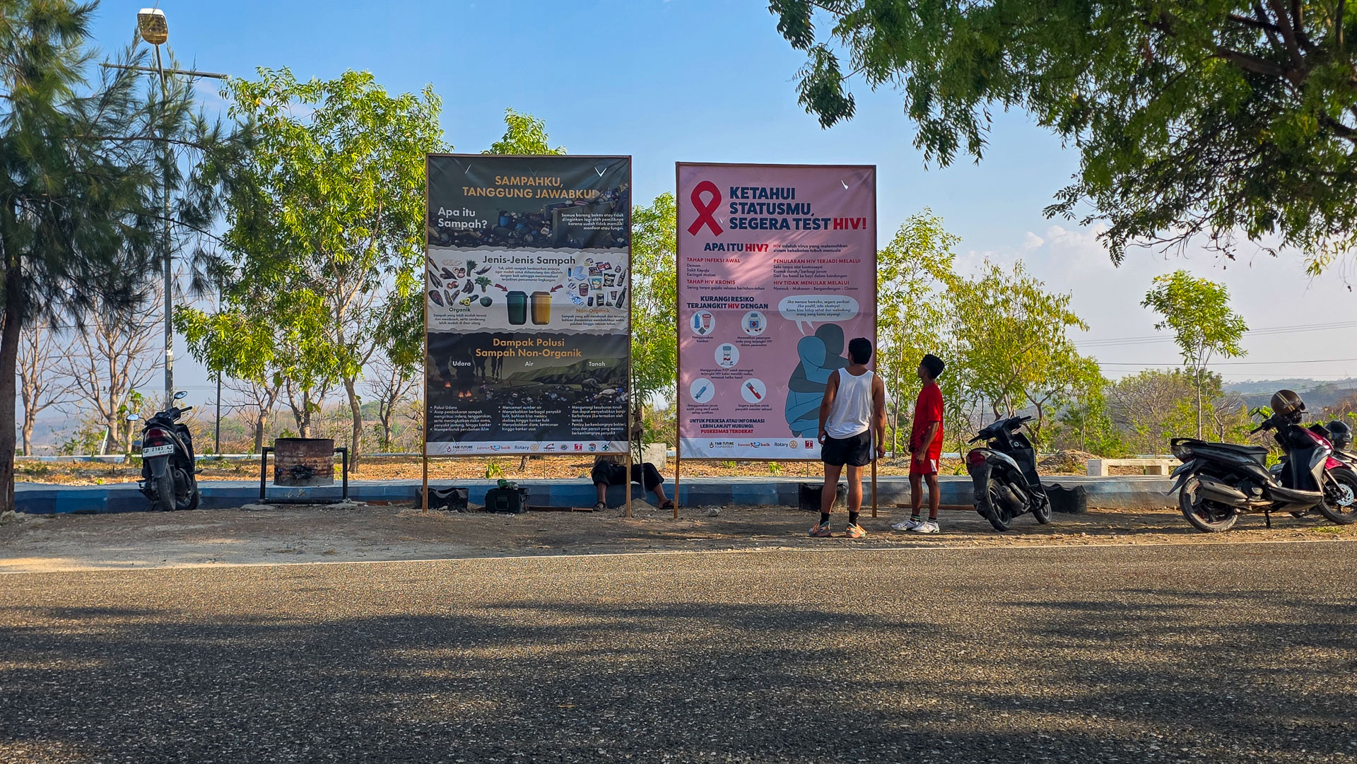 Community health education beside Waingapu’s jogging trail People stand beside HIV and waste billboards near parked scooters and trees in Waingapu.