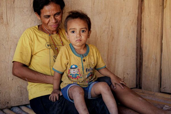Mother in a yellow shirt holding a young child on her lap beside a wooden wall in Napu, East Sumba.