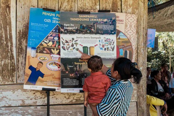 Mother holding her child beside waste, nutrition and malaria education posters in a remote East Sumba village.