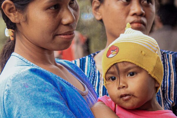 A woman in a blue top holds a young child wearing a yellow knitted hat, with another woman behind them.