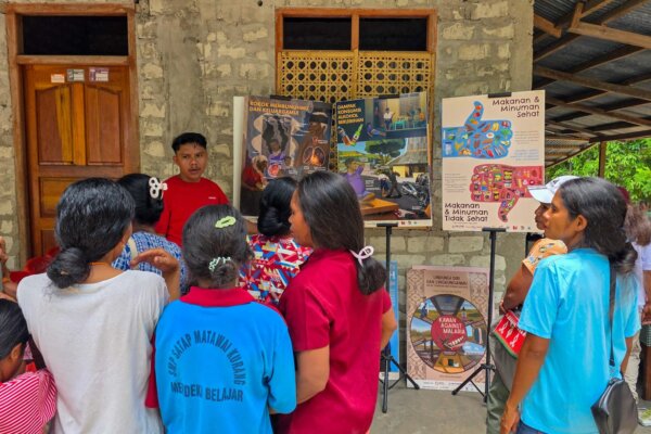 Erwin in red speaks to a group beside tobacco, alcohol and nutrition posters, with a malaria poster below.
