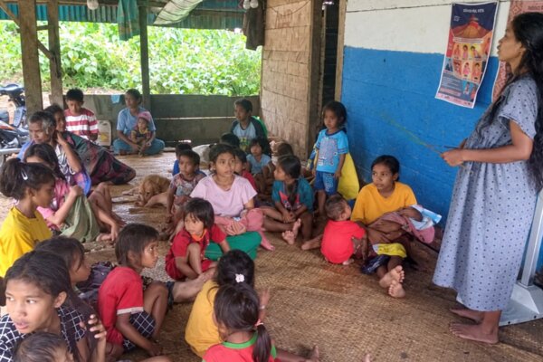 Children and adults on woven mats beside a woman using a pointer and health education posters in Laihiru.