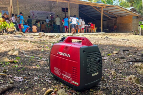 Rotes Honda EU22i-Generator auf dem Boden vor einem Gebäude, in dem Familien in Ost-Sumba für die Gemeinschaftsversorgung zusammenkommen.
